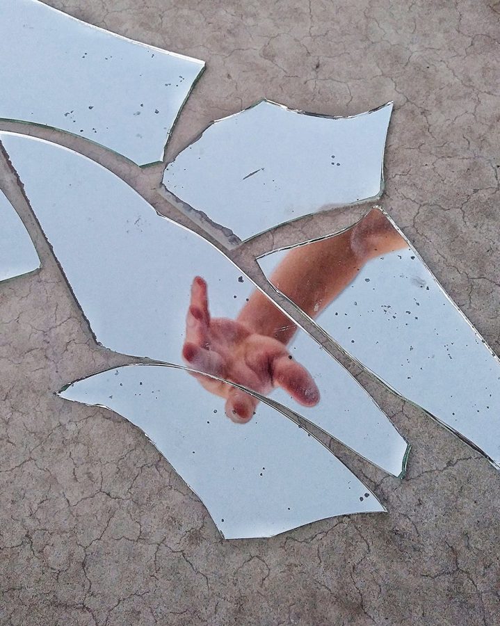A photograph of glass shards laying across the ground with a hand reaching out toward them in the reflection.
