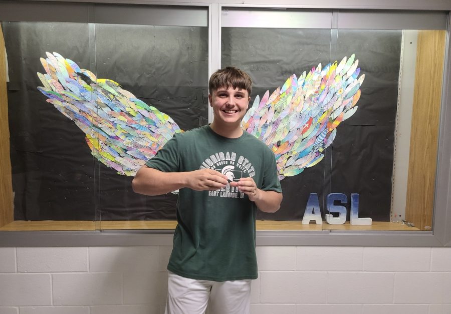 Senior Carter Lyons standing in front of an ASL project signing his name sign. 